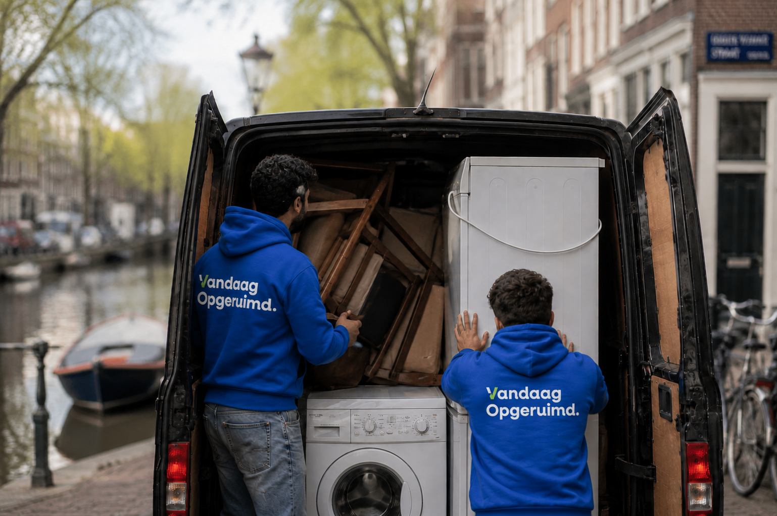 Twee medewerkers in Vandaag Opgeruimd hoodies laden een koelkast en wasmachine in een bus langs een Amsterdamse gracht