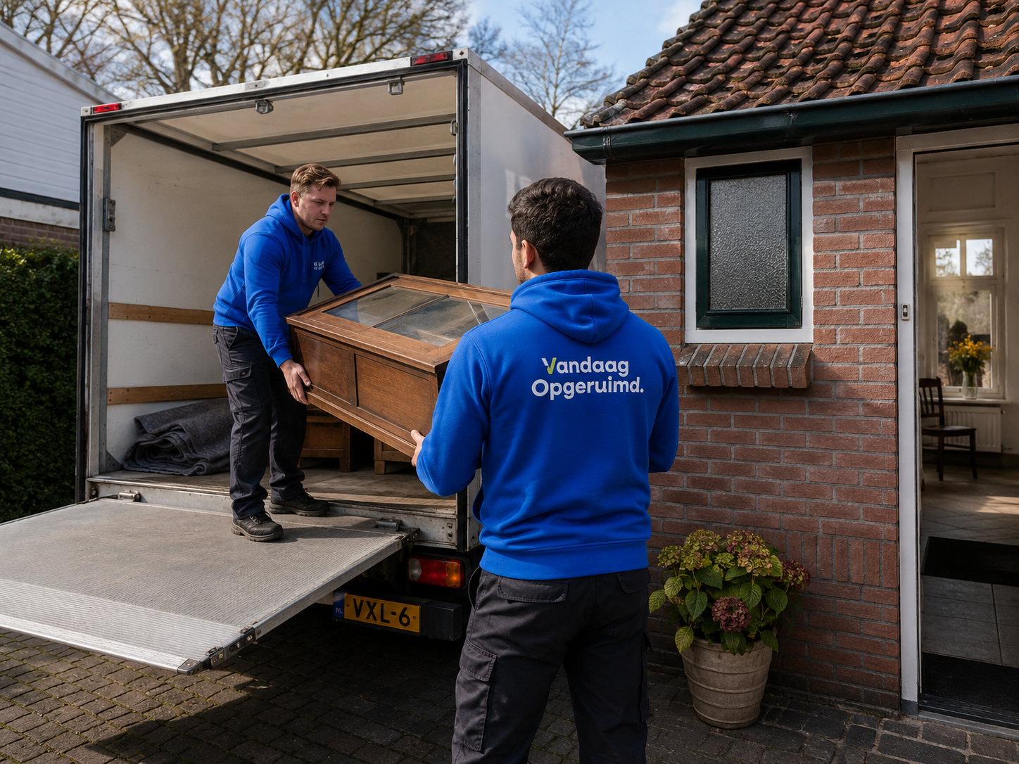 Twee medewerkers in Vandaag Opgeruimd hoodies laden een kast in een ophaalbus bij een woning